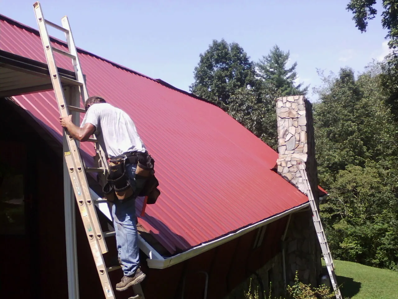 Roofer on a ladder beside a red metal roof and stone chimney — Soffit & Fascia Repair by Roofing Brothers in Sylmar